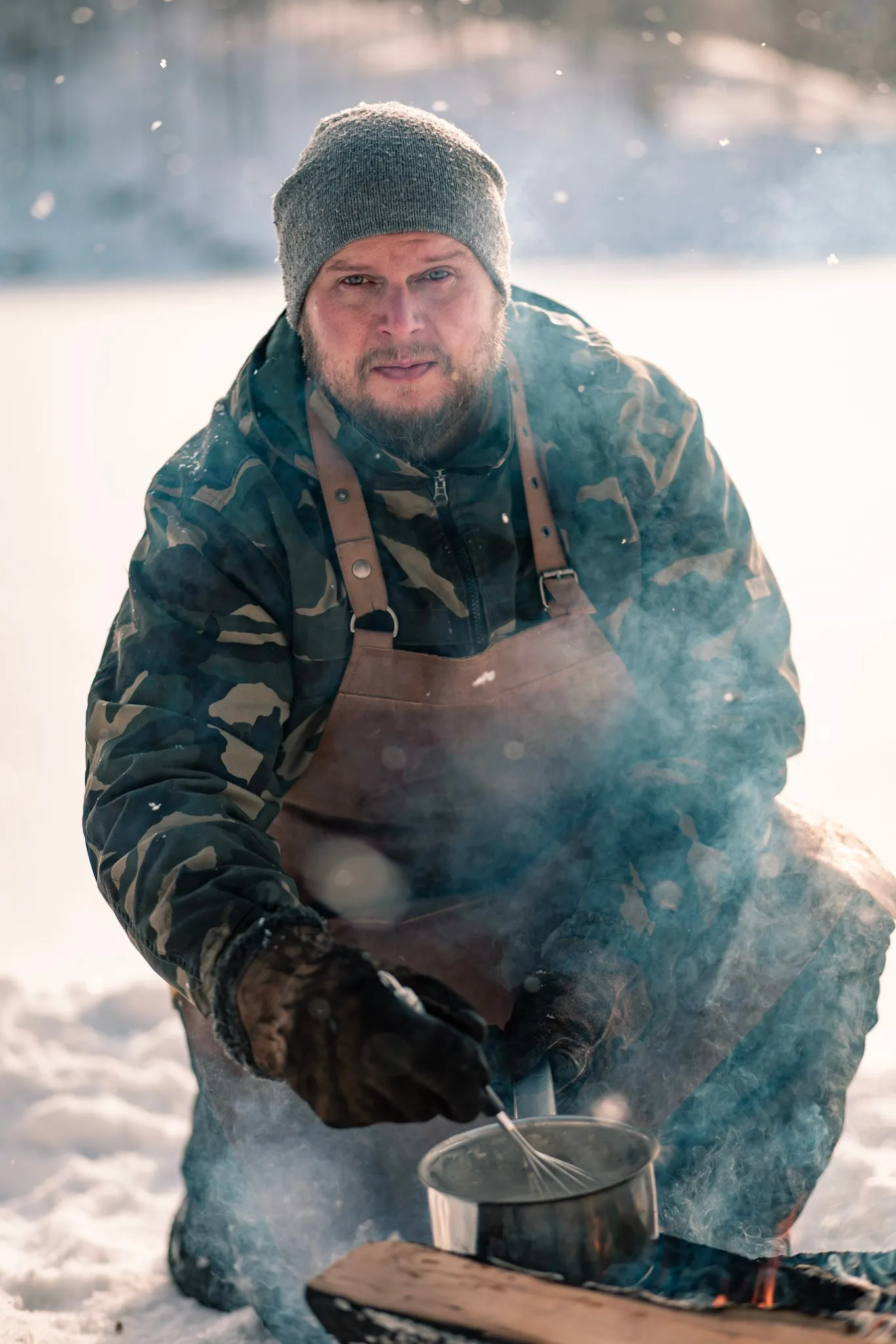 Mies talviulkoilmassa työskentelemässä, sekoittaa kattilaa avotulen äärellä lumisessa ympäristössä.